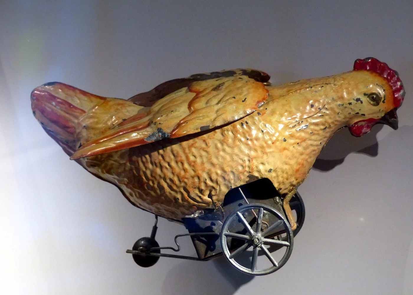 Tin toy hen of about 1880 painted in shaded browns and red, its body mounted on a small wheeled frame with a clockwork mechanism visible beneath