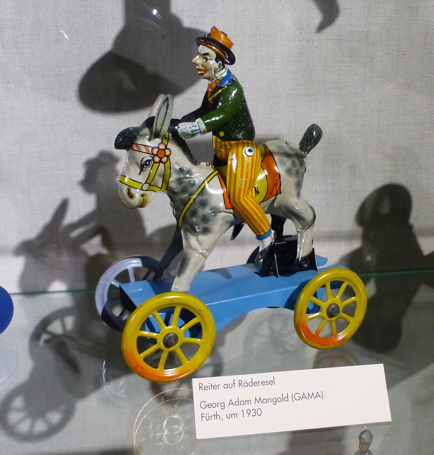 Tin toy of a rider on a wheeled donkey, brightly lithographed in blue, yellow and red, displayed in a museum case beside a printed label giving maker, town and date