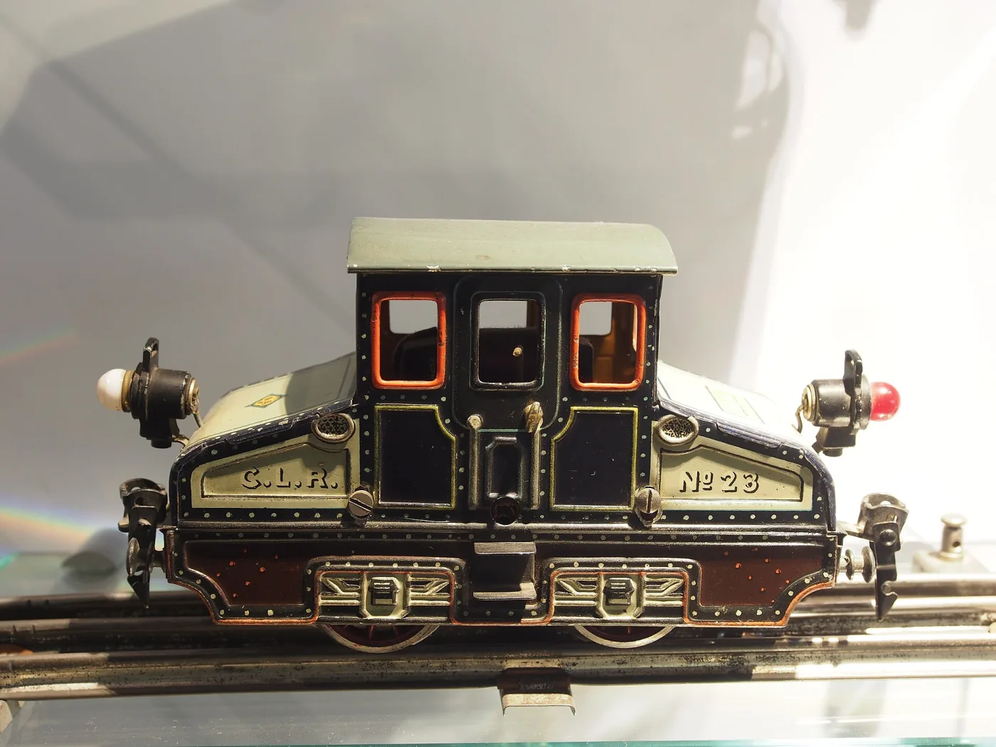 Tin toy locomotive of about 1910 in dark green and black with red-lined running boards, brass fittings and a tall chimney, standing on a length of track in a museum case