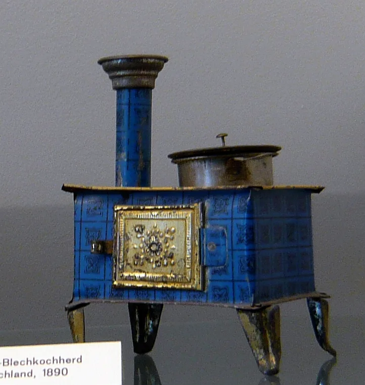 Small tin toy cooking stove printed in blue with an ornamental panel on its door, a chimney at one side and a lid on top, standing on four short legs beside a museum label