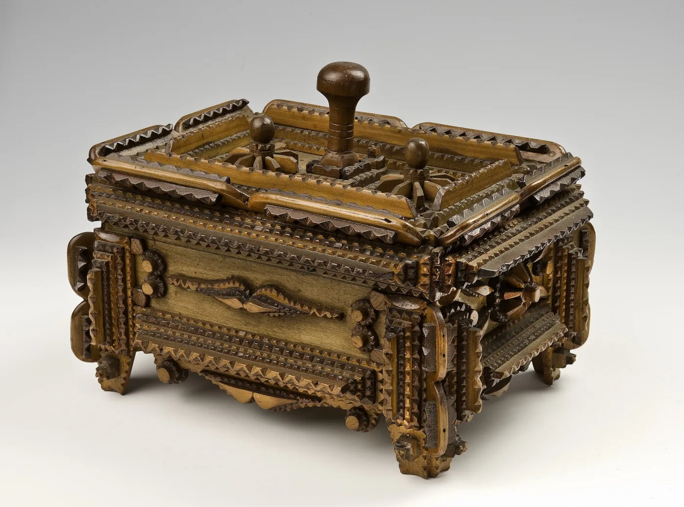 Two-tone tramp art casket on carved feet, its stepped lid and corner buttresses built from many notched tiers of alternating pale and dark wood, with three turned knobs on top