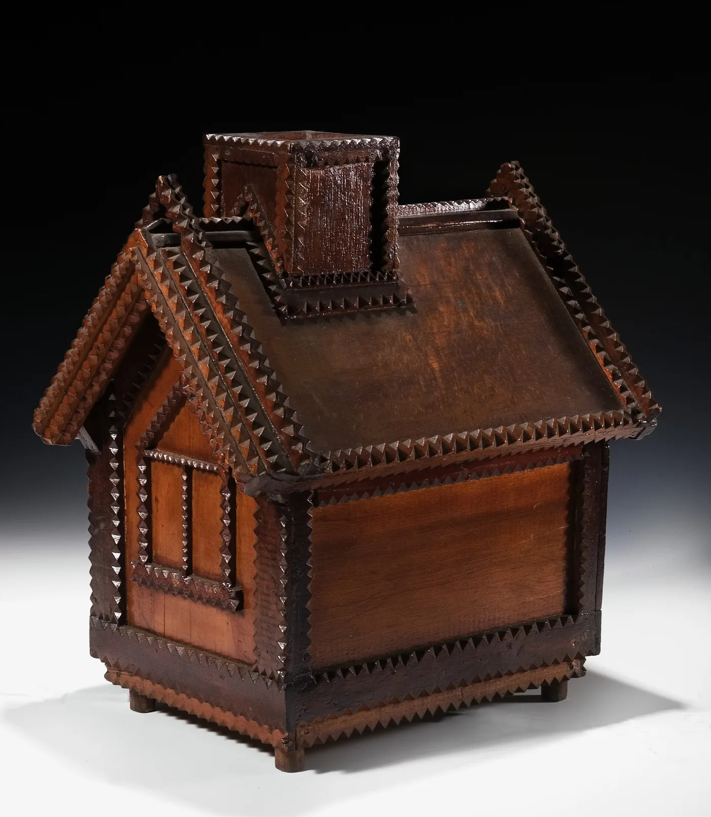 Tramp art box built as a gabled house with a chimney, its roof and wall panels in pale plain wood and every edge outlined by dark notch-carved strips