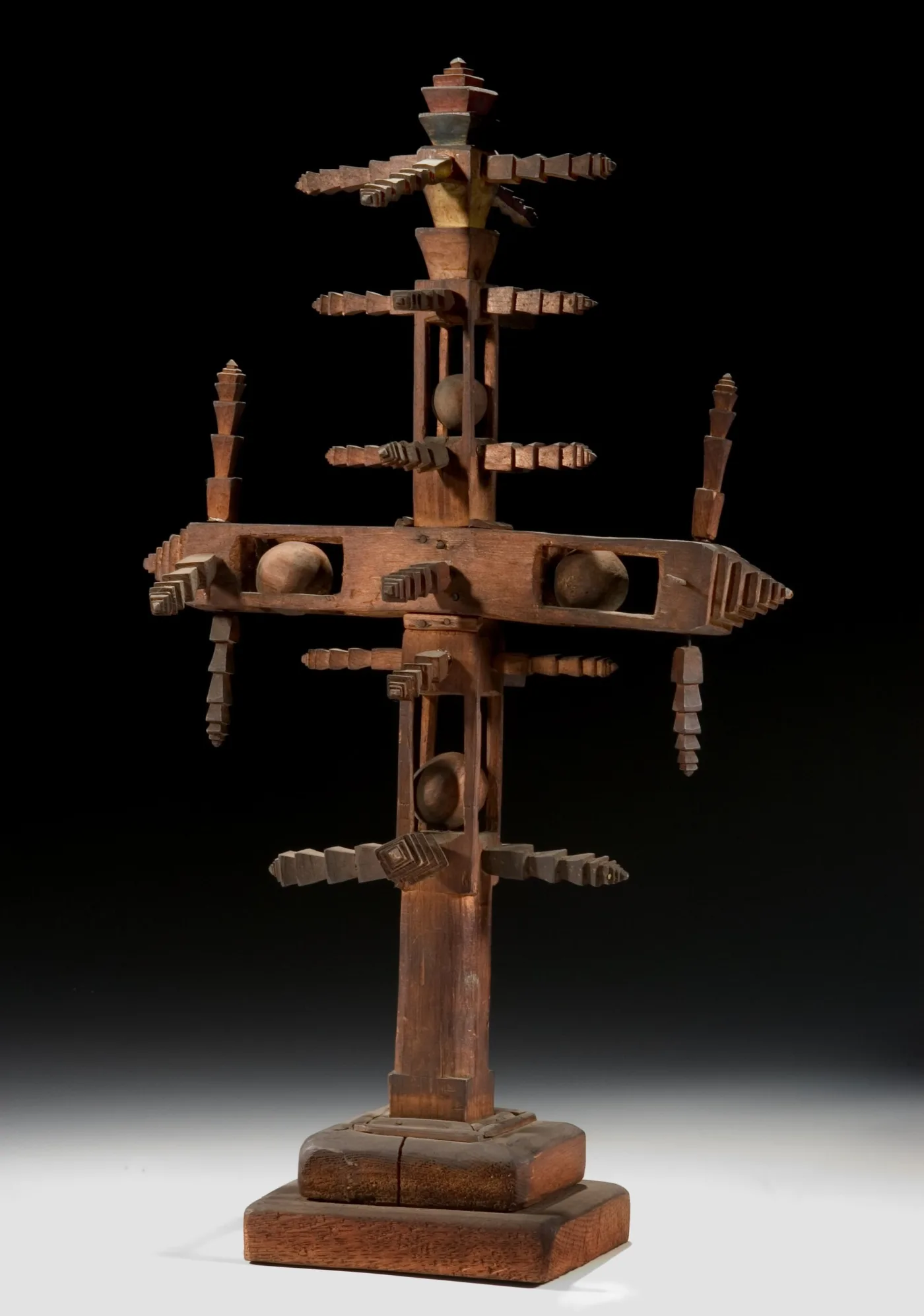 Cruciform wooden object on a stepped base, its arms formed from stepped notched strips, with loose carved balls trapped inside open cage sections of the shaft