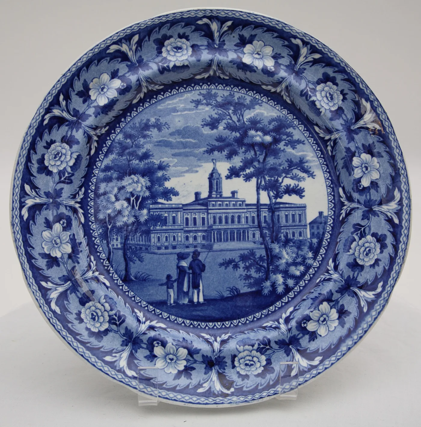 Blue and white plate showing a classical marble civic building behind railings with figures in the foreground, inside a wide border of flowers and foliage