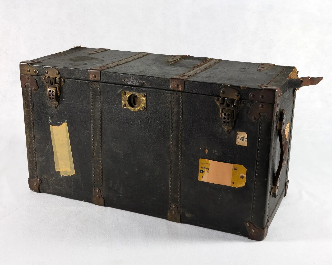 Black canvas-covered flat-topped travel trunk with metal edge binding and brass catches, its lid and sides carrying pale paper labels and stickers
