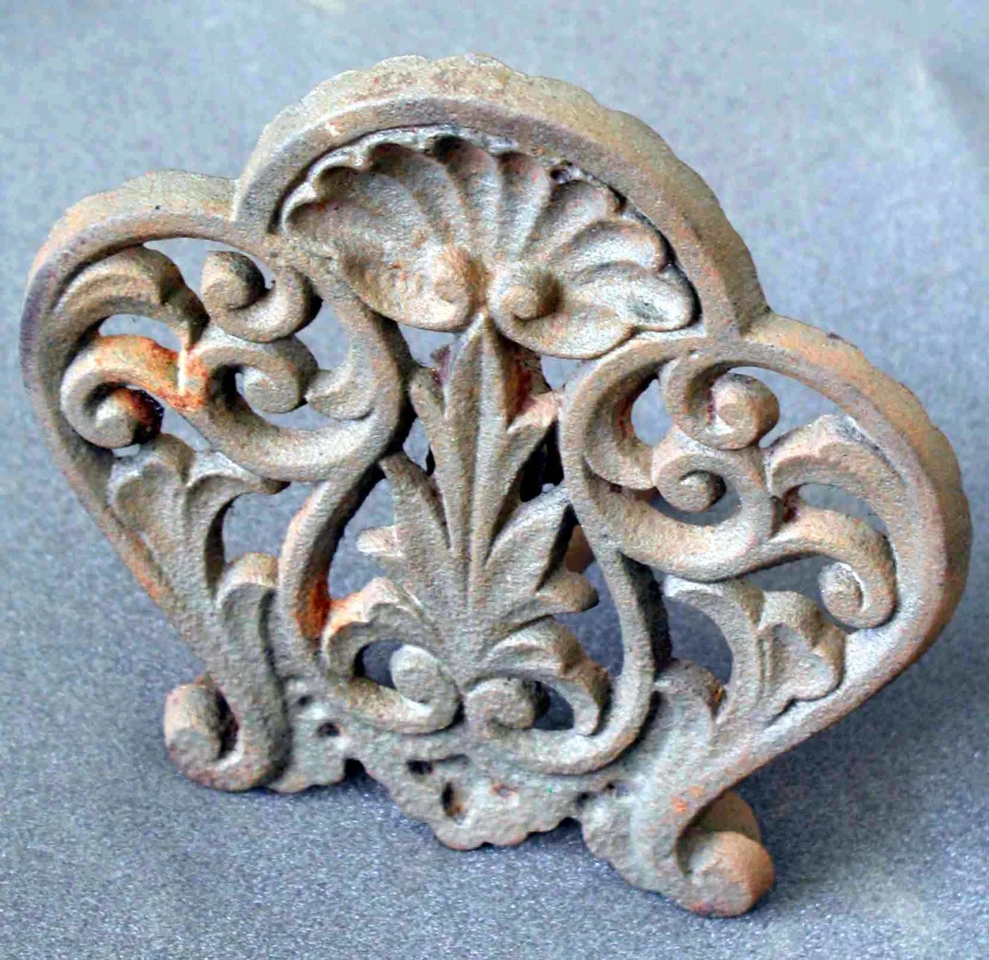Heart-shaped cast iron trivet with a scallop shell at the top and scrolling foliage filling the interior, rusted overall, photographed at an angle