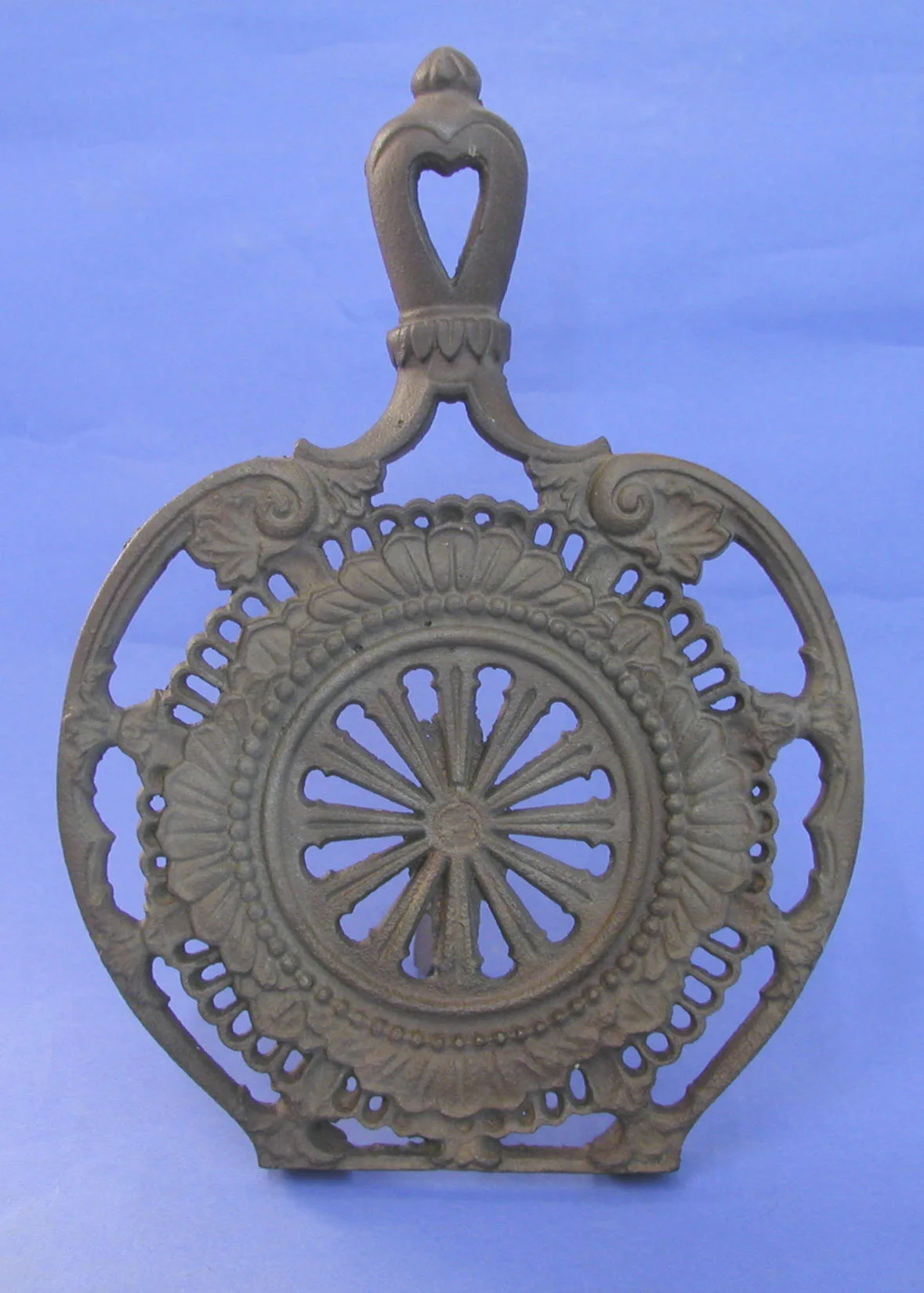 Cast iron trivet seen from the front: a spoked wheel rosette inside beaded rings with fan-shaped foliage around it, and a handle pierced with a heart