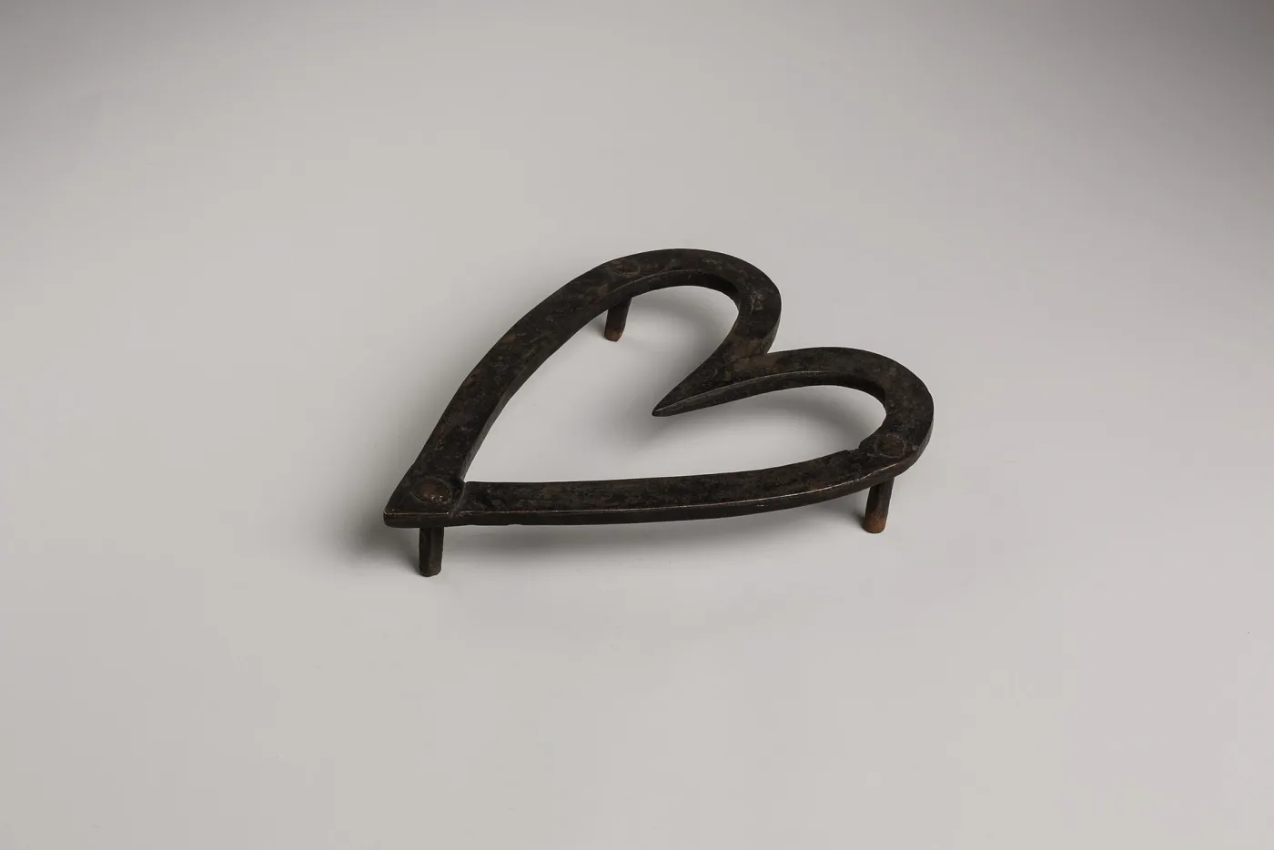 Heart-shaped forged iron trivet raised on three short cylindrical feet, photographed on a plain grey ground