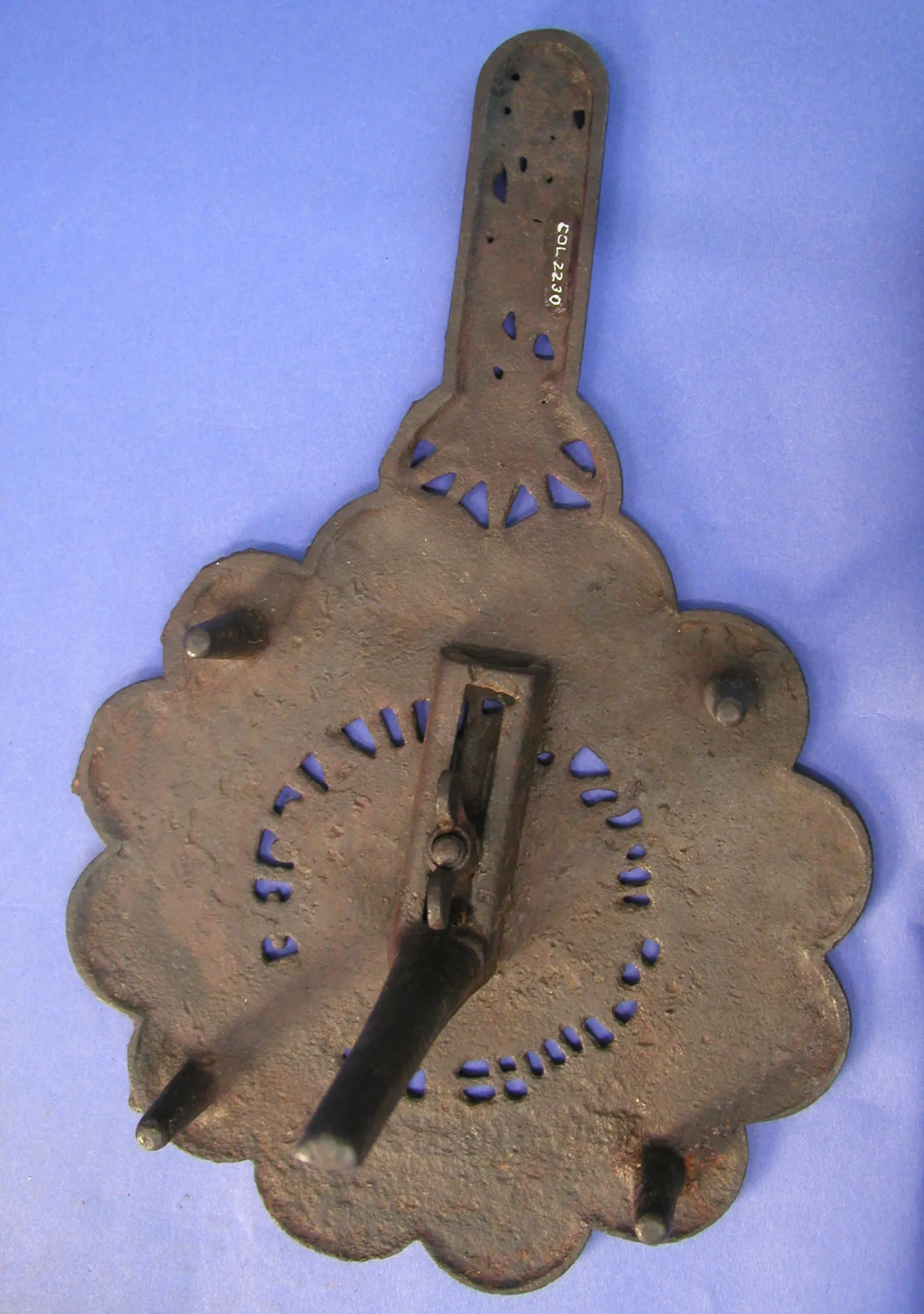 Underside of a scalloped cast iron trivet showing four short tapering feet growing straight out of the body, and a hinged iron bar riveted across the centre
