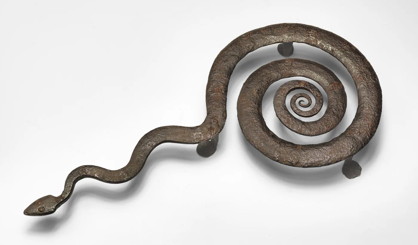 Wrought iron trivet formed from a single snake, its coiled body making the round top and its tail extending sideways as the handle, on three stub feet