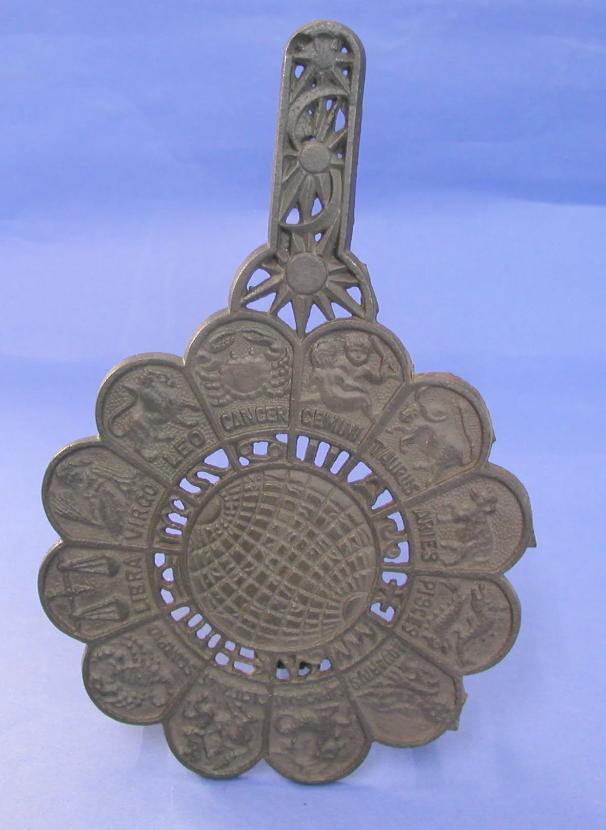 Scalloped cast iron trivet, each of its twelve lobes carrying a named sign of the zodiac in relief around a pierced gridded globe, with a pierced sunburst handle