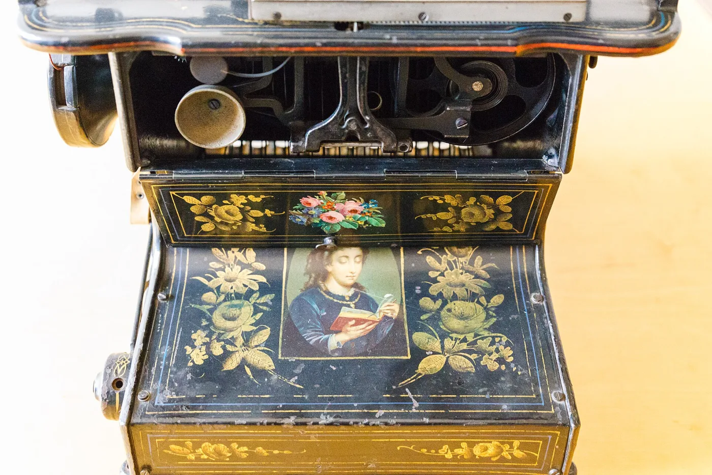 Close view of the decorated top of a Sholes and Glidden typewriter, its black japanned surface painted with a landscape panel surrounded by gilt scrollwork and sprays of coloured flowers