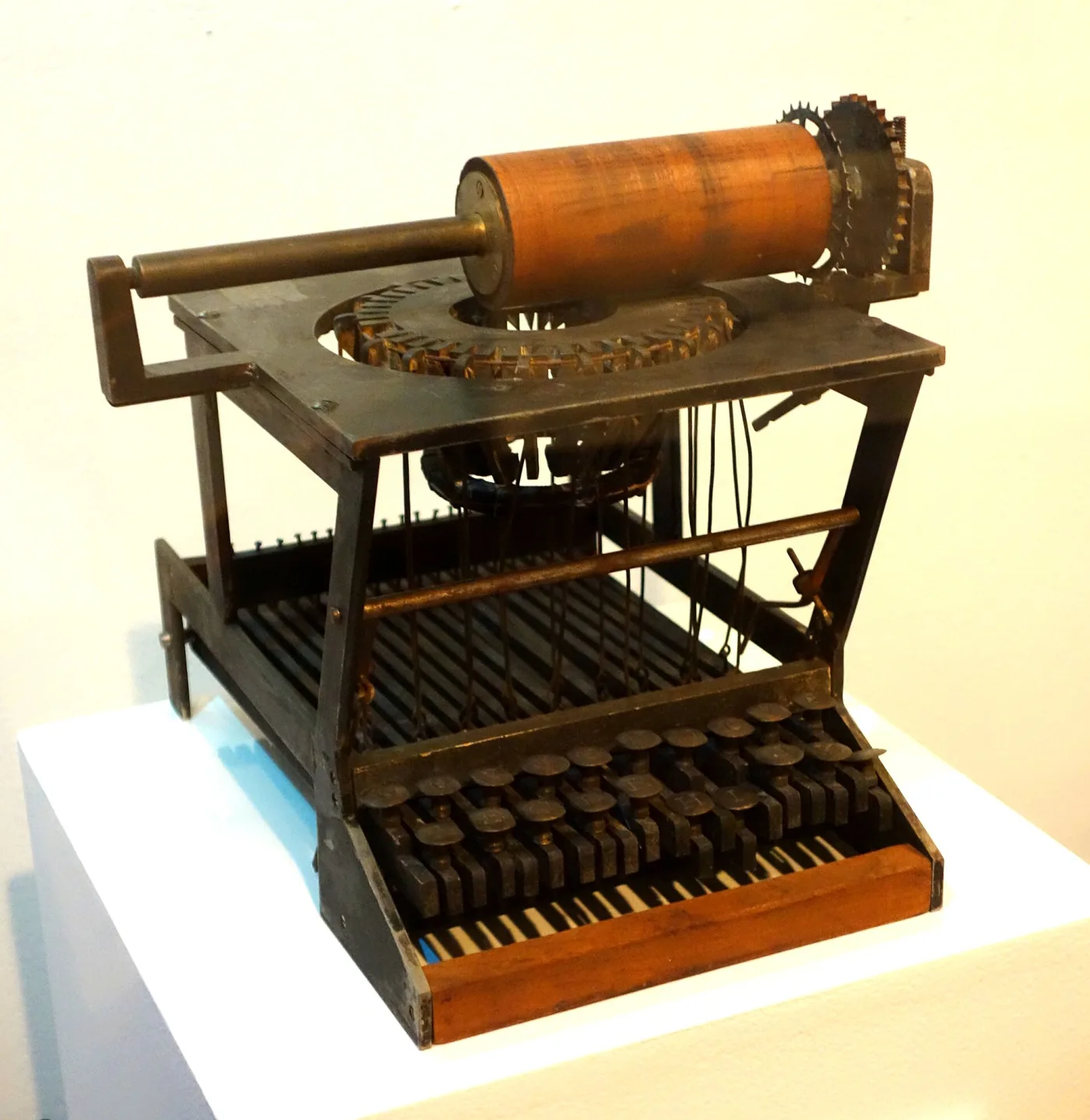 Skeletal early typewriter mechanism with an open wooden frame, a brass cylinder platen above and rows of exposed type levers below, displayed on a plain plinth
