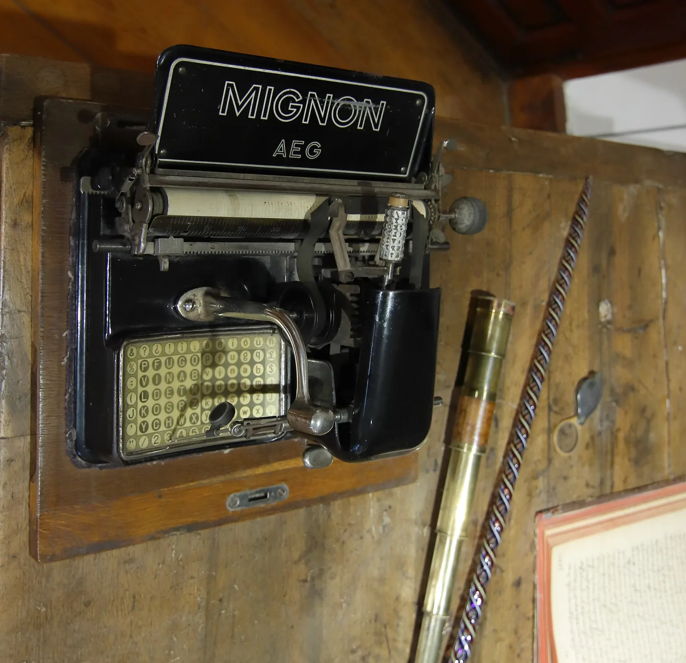 AEG Mignon index typewriter on a wooden base, with an upright printed character chart on the left, a pointer arm resting on it, a small cylindrical type sleeve at the centre and a single key at the right