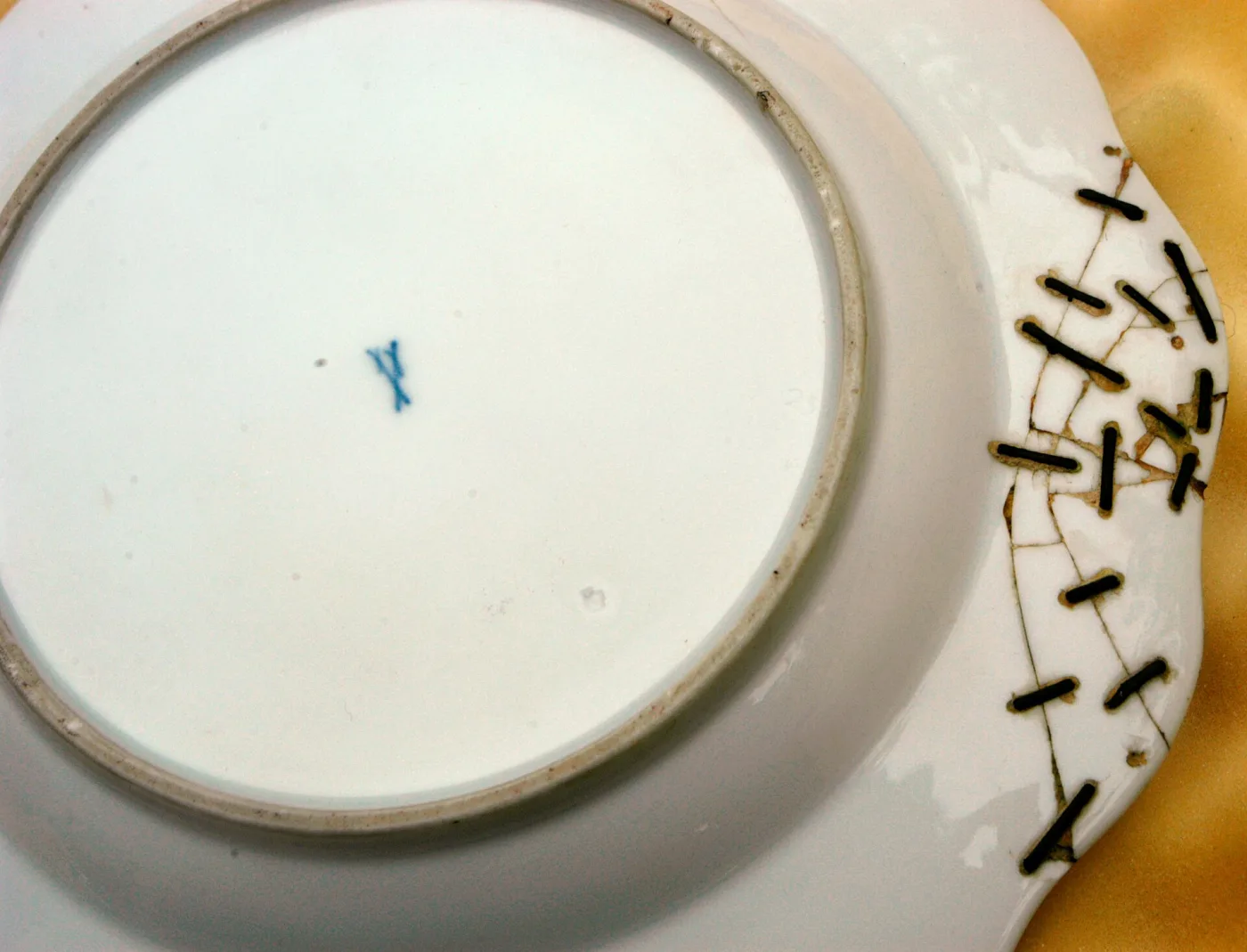 Underside of a white porcelain plate showing a small blue factory mark at the centre and about fifteen iron staples set in drilled holes across a repaired break in the rim