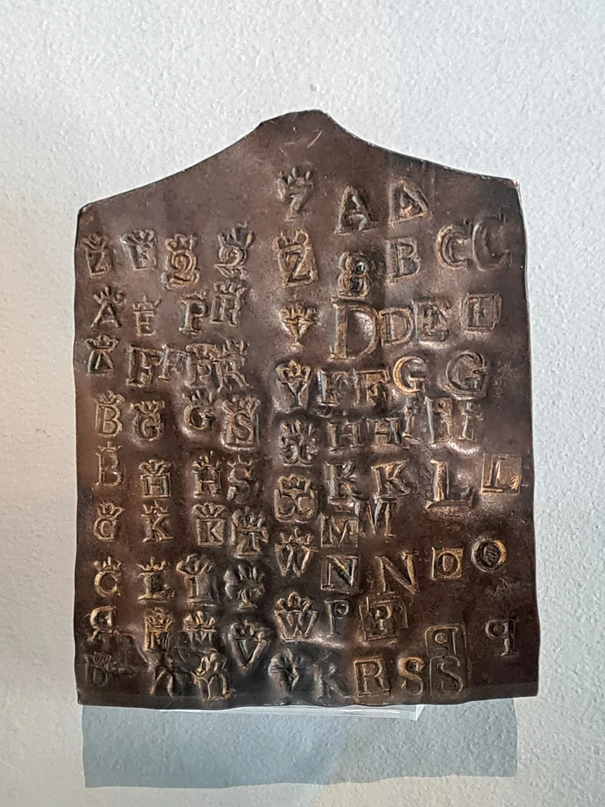 Dark copper plate covered with dozens of small struck letter punches, most surmounted by a crown, mounted against a pale wall