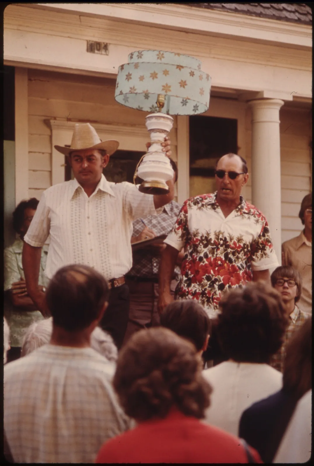 Colour photograph of a man in a straw hat holding a table lamp above his head while an auctioneer calls bids and a crowd watches from a lawn