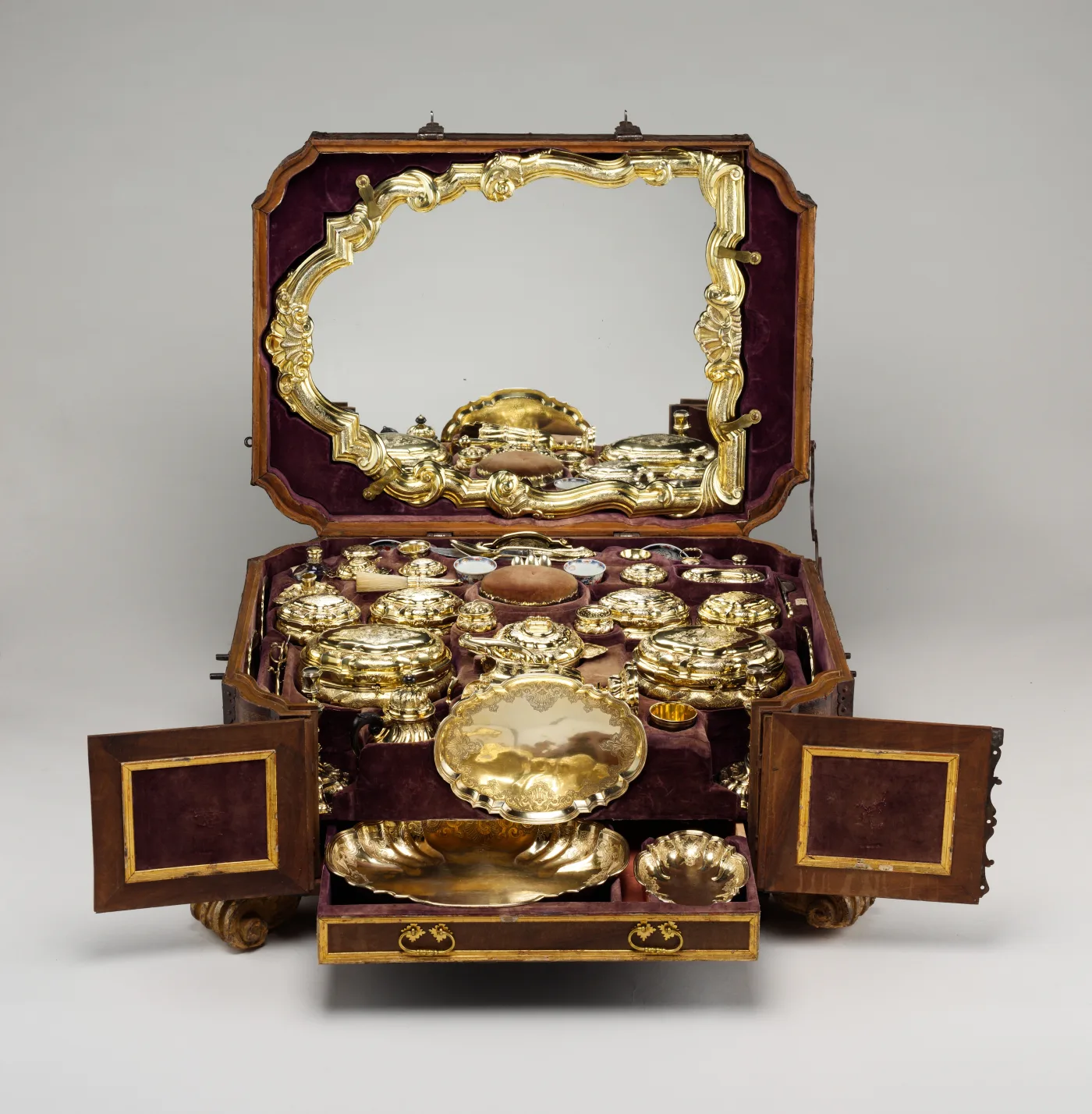 Open leather travelling case lined in purple velvet, a rococo silver-gilt mirror standing in the lid and the base packed with fitted silver-gilt covered boxes, a salver, porcelain cups and a pin cushion
