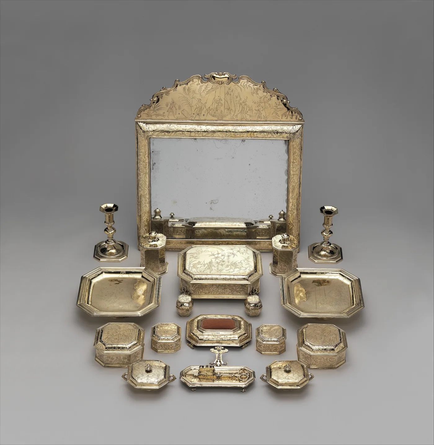 Silver-gilt toilet service laid out on grey: a large mirror with a shaped cresting engraved with chinoiserie figures, flanked by candlesticks, with salvers, caskets, covered boxes and a pin cushion arranged in front