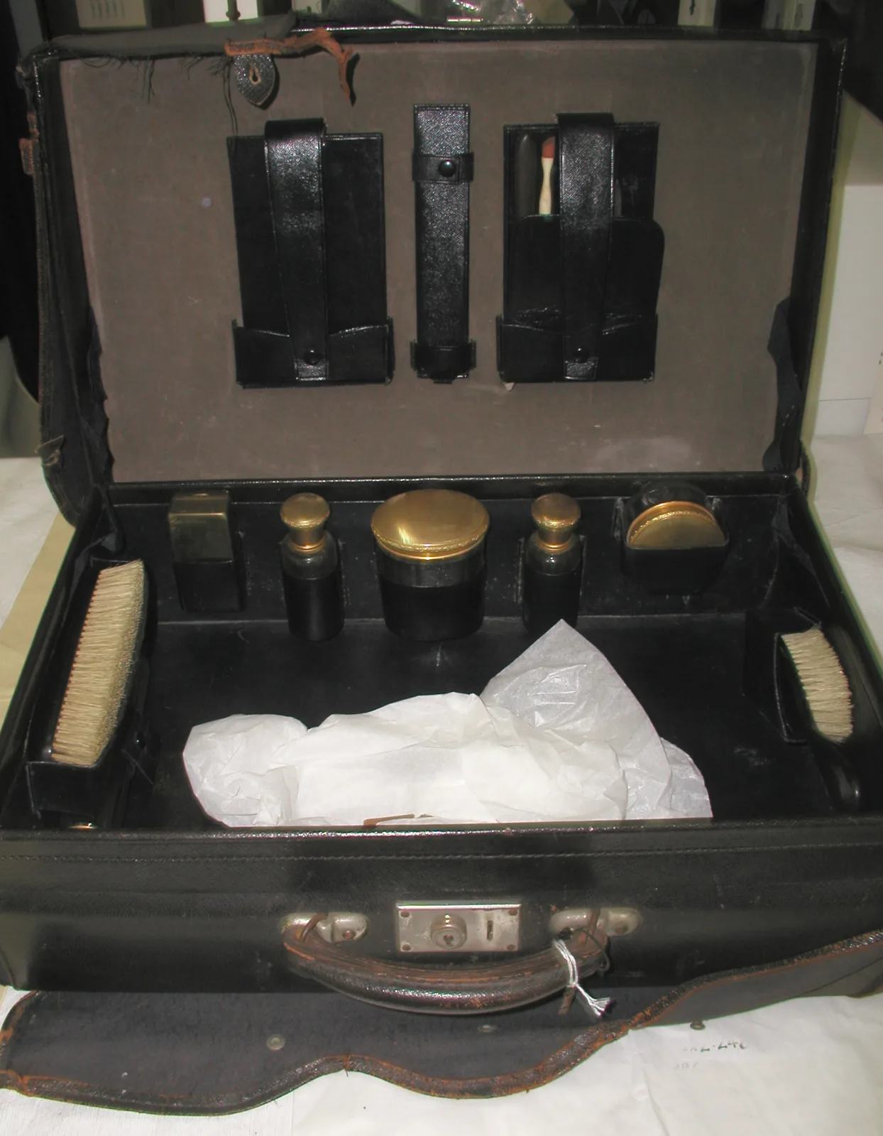 Open black leather dressing case with gilt-topped glass jars and bottles seated in fitted wells, two clothes brushes at the sides, leather loops in the lid, and empty spaces where items are missing