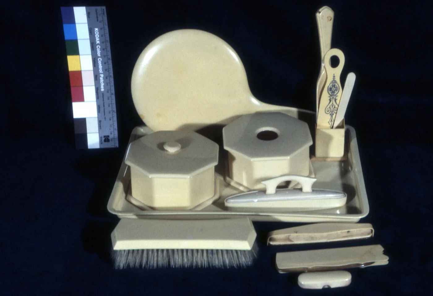 The same celluloid set seen from the other side, showing the plain back of the hand mirror, a bristle clothes brush, a buffer and small tools, one piece brown-stained and split, against a dark ground
