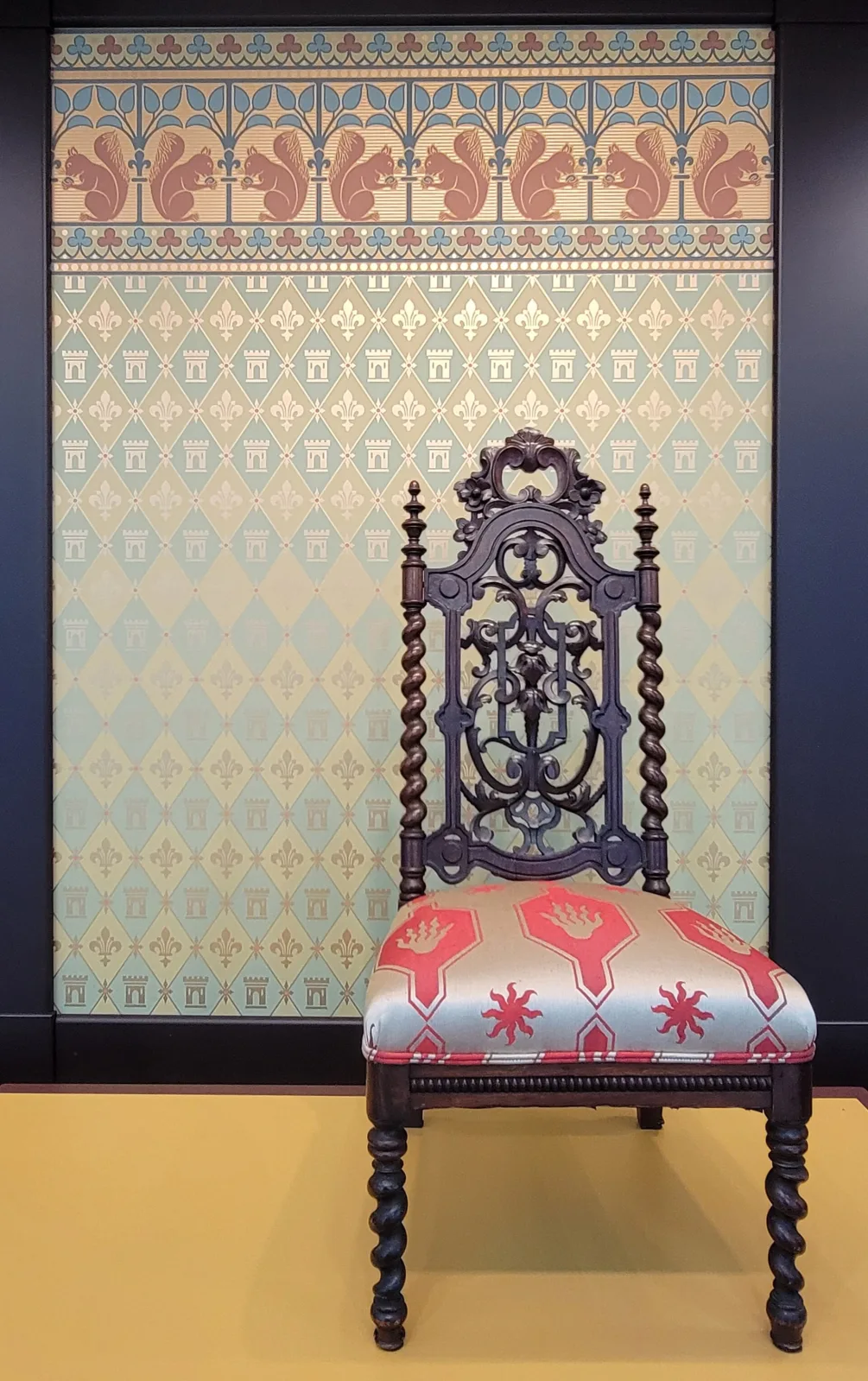 Tall dark hall chair with barley-twist uprights and legs and a pierced scrolled back, standing against patterned wallpaper of fleur-de-lys, castles and squirrels