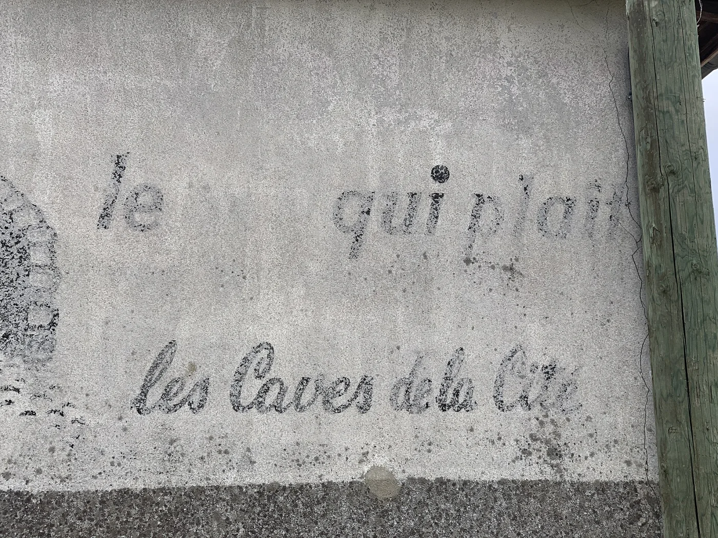 Faded painted advertisement on a rendered farmhouse wall, only fragments of cursive lettering still legible above the words les Caves de la Cite