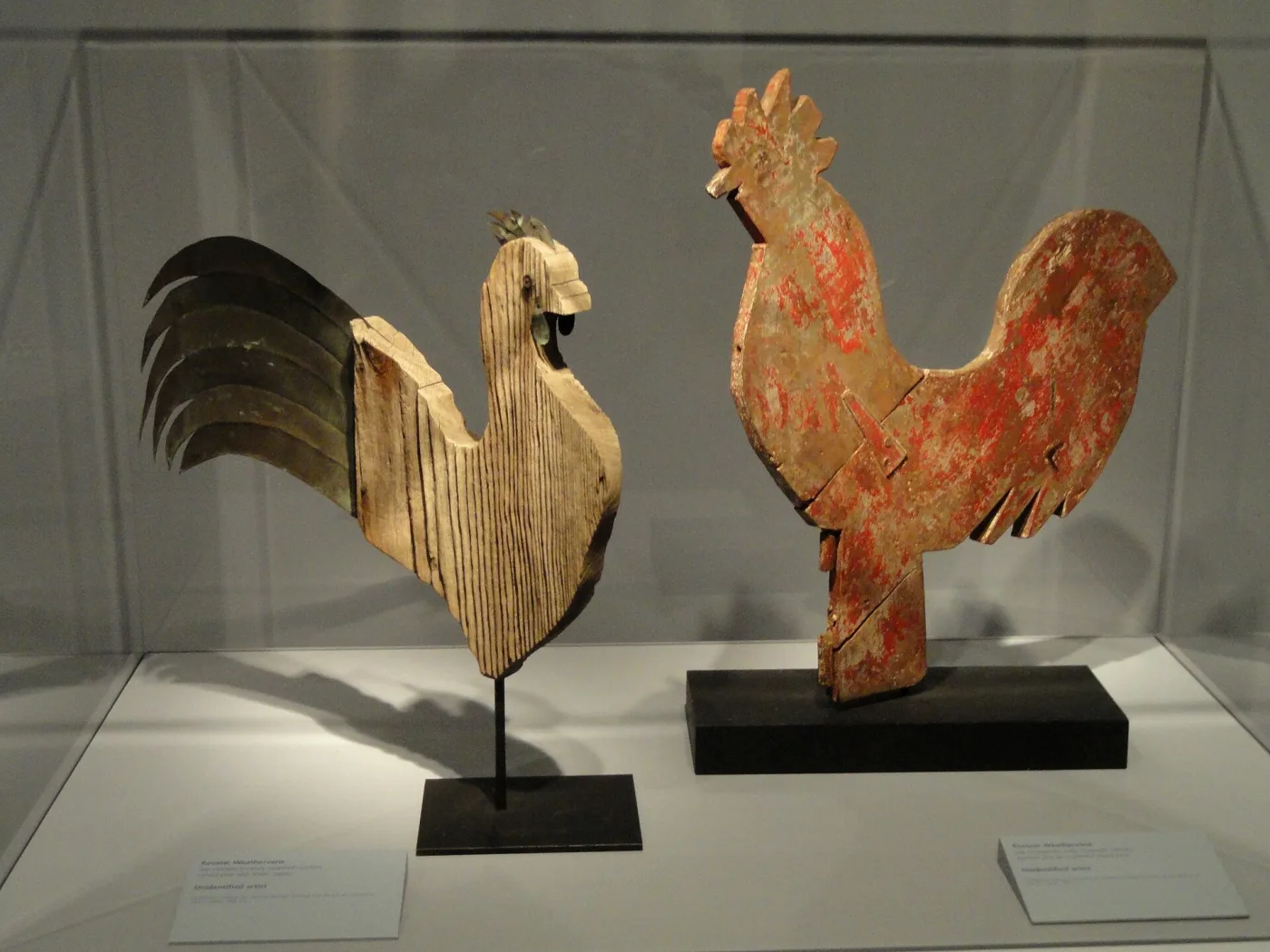 Three copper rooster weathervanes of differing designs displayed together on stands in a museum gallery