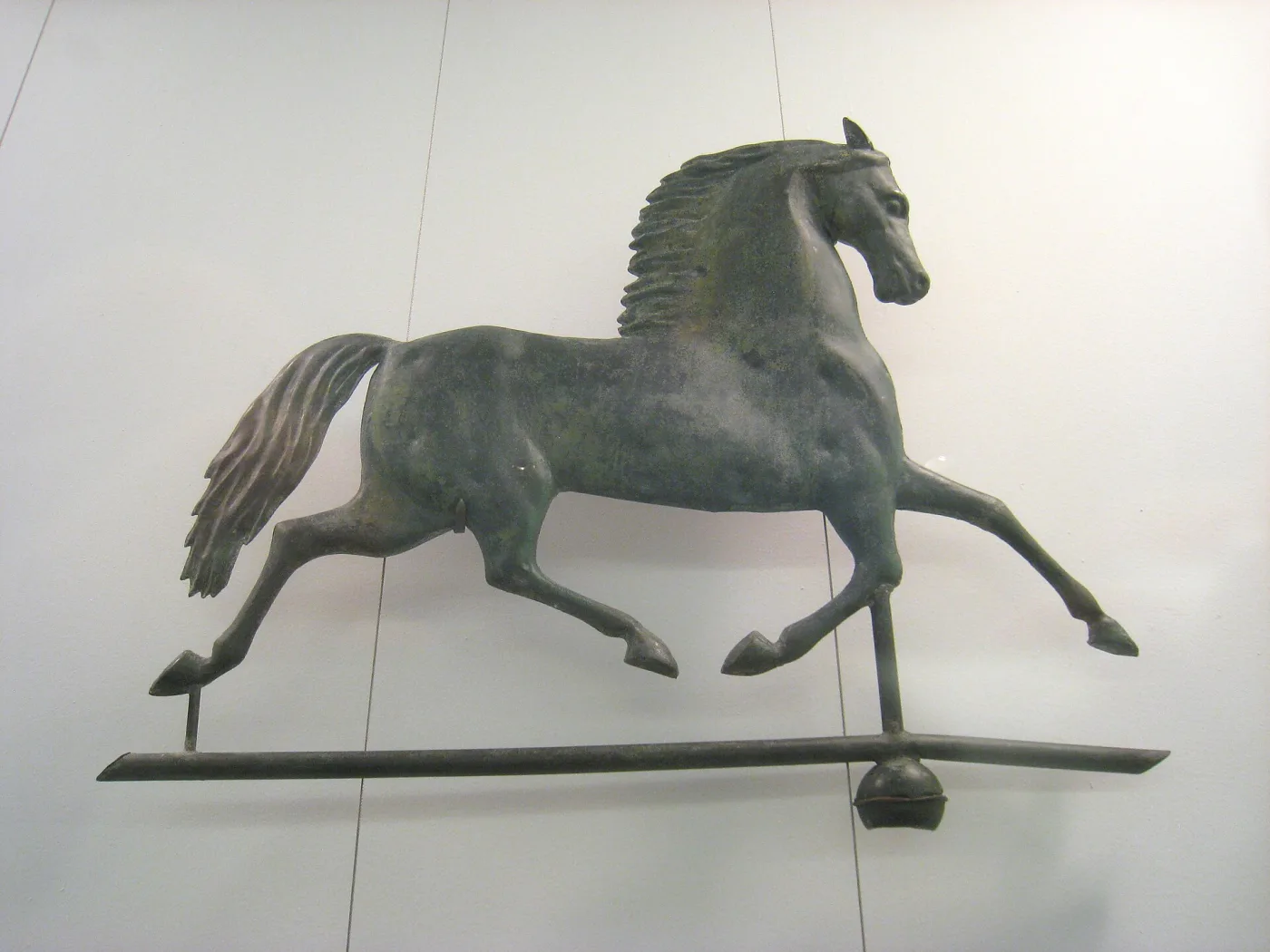 A copper trotting horse weathervane with a warm brown and green surface, shown on a stand in a museum case