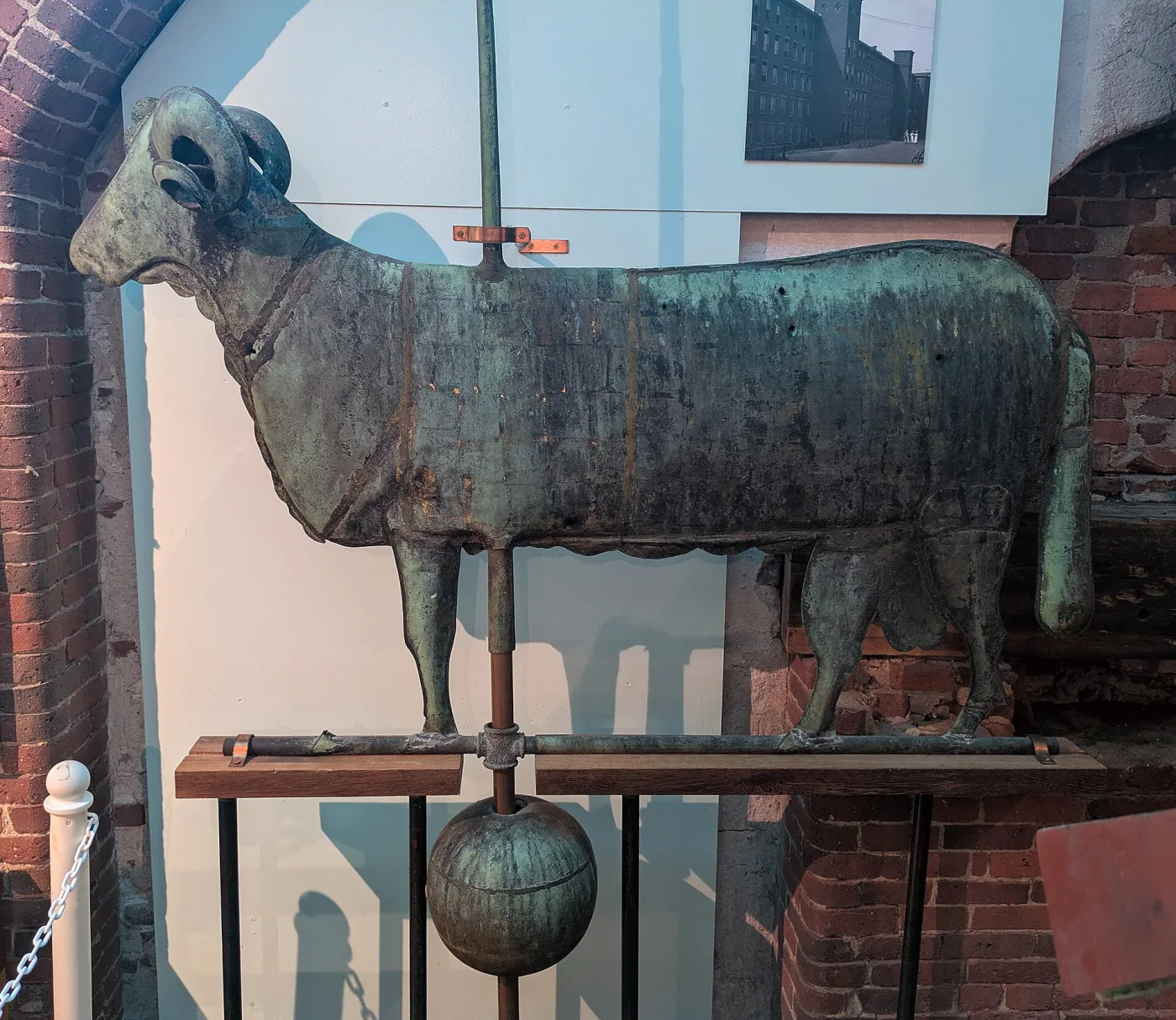 A large copper ram weathervane covered in green verdigris, with a visible vertical seam along its body, several small holes, and a directional rod with a copper ball beneath it