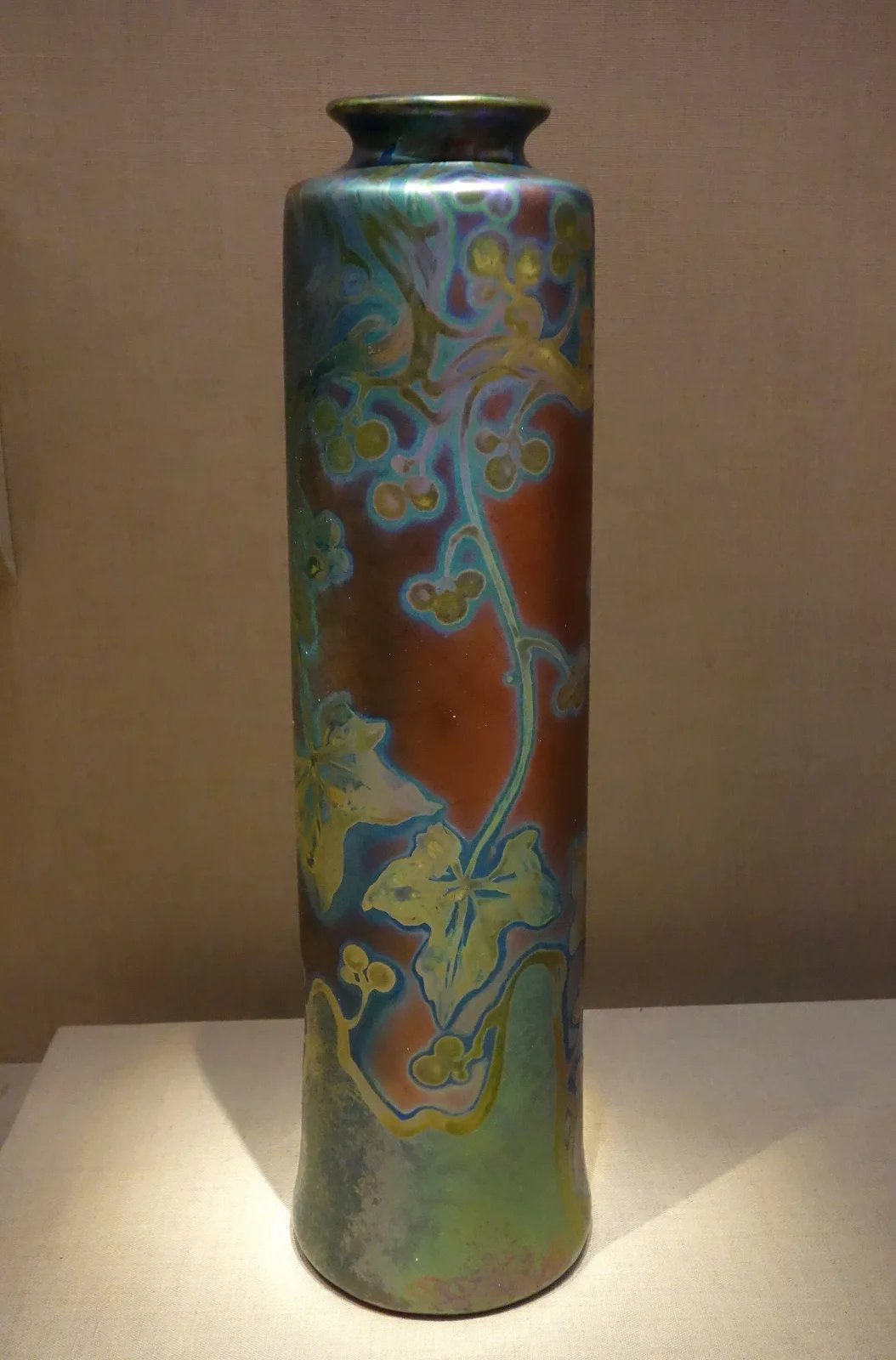 Tall cylindrical vase with a metallic iridescent glaze shading from red-gold to green and blue, decorated with sinuous trailing stems and leaves that catch the light differently from the ground
