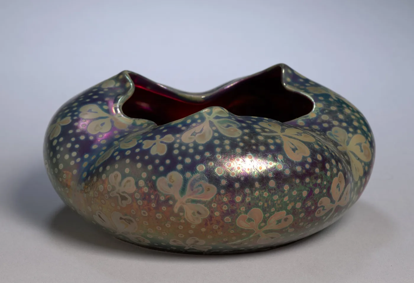 Low squat bowl with an irregular pinched rim, its iridescent surface scattered with pale flower heads over a mottled purple, gold and green lustre, the interior glazed deep red