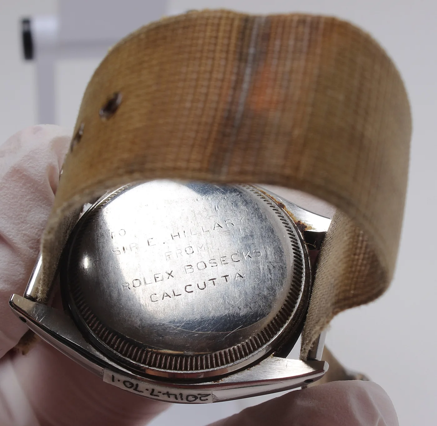 Gloved hands holding a wristwatch with a woven fabric strap, showing a case back engraved To Sir E. Hillary from Rolex Bosecks Calcutta