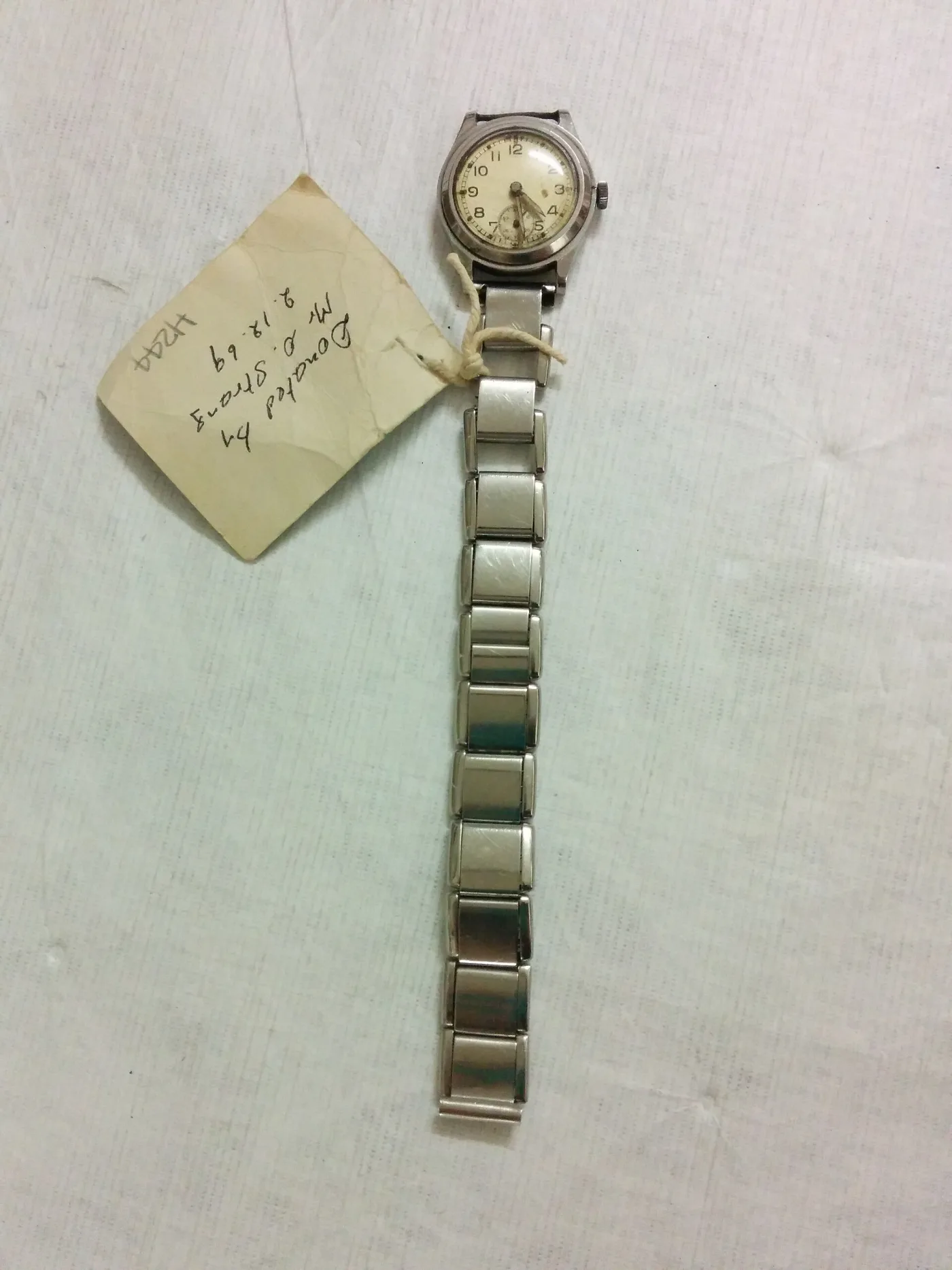 Small round wristwatch with a cream dial and subsidiary seconds on a flexible expanding metal bracelet, beside a handwritten museum donor tag