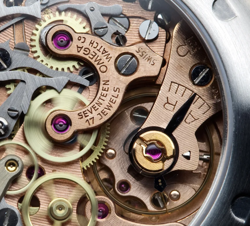 Close-up of a copper-coloured watch movement bridge engraved Omega Watch Co, Seventeen 17 Jewels, Swiss, with ruby jewels set into it