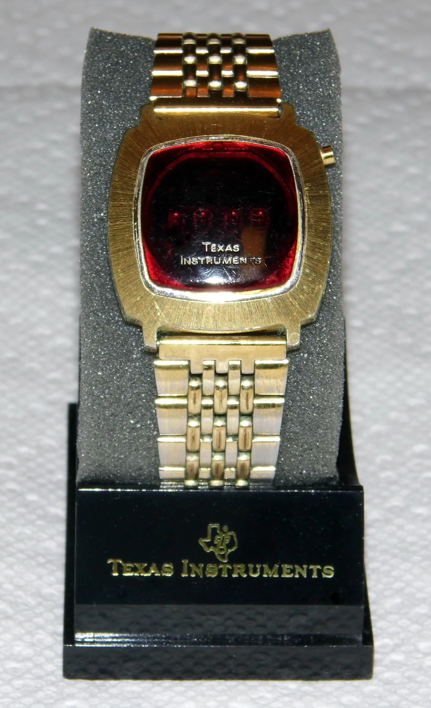 Gold-tone digital wristwatch with a dark red display window marked Texas Instruments, on a branded retail display stand
