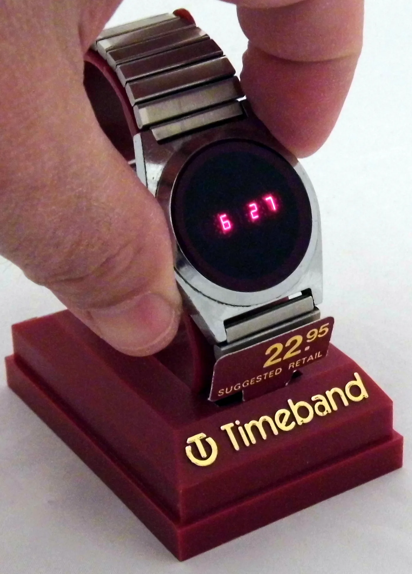 Steel digital wristwatch showing 6:27 in red LED digits, held above a shop display base marked Timeband with a 22.95 dollar price tag