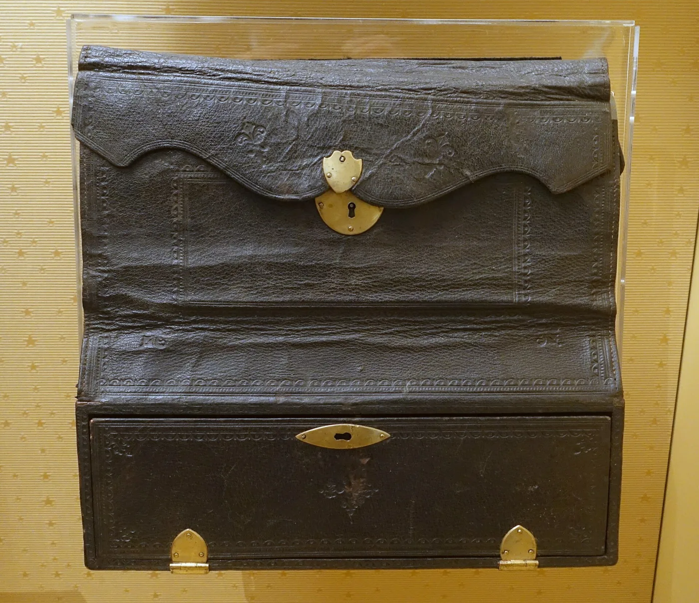 Leather-covered travelling writing case with a shaped flap fastening over the front, a small escutcheon at the centre and pale wooden feet, standing on a yellow museum plinth