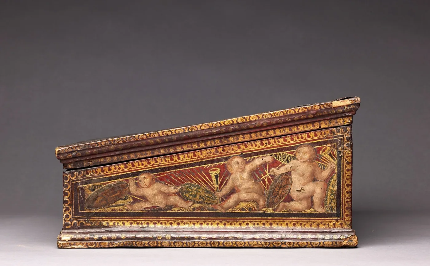 Front of a sixteenth-century writing box painted and gilded with winged putti flanking a central roundel, above a band of small painted panels along the base moulding