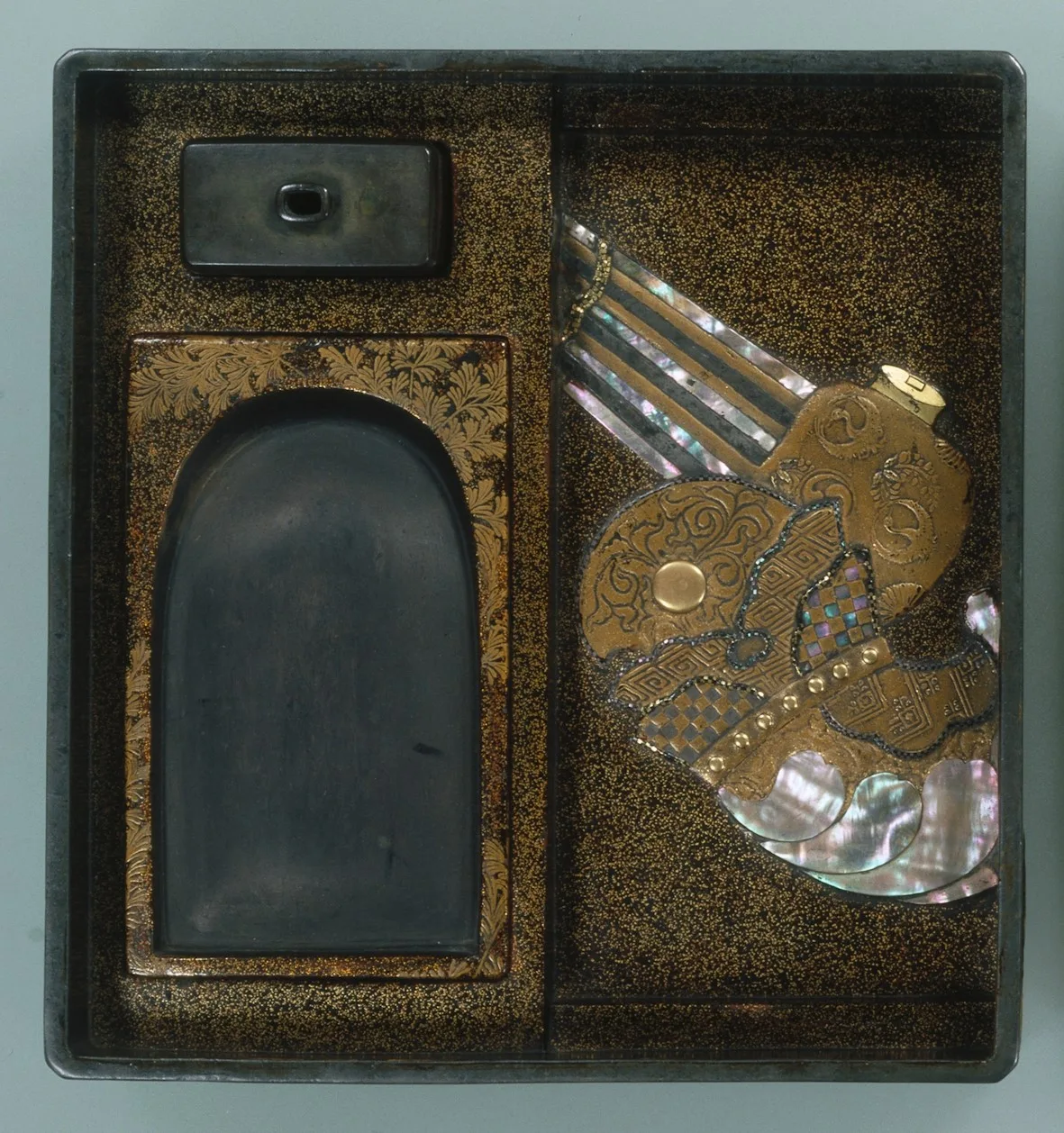 Open lacquer writing box showing its fitted interior: a black rectangular inkstone in a recess, a metal water dropper beside it, and shaped compartments holding brushes and a mother-of-pearl decorated tray