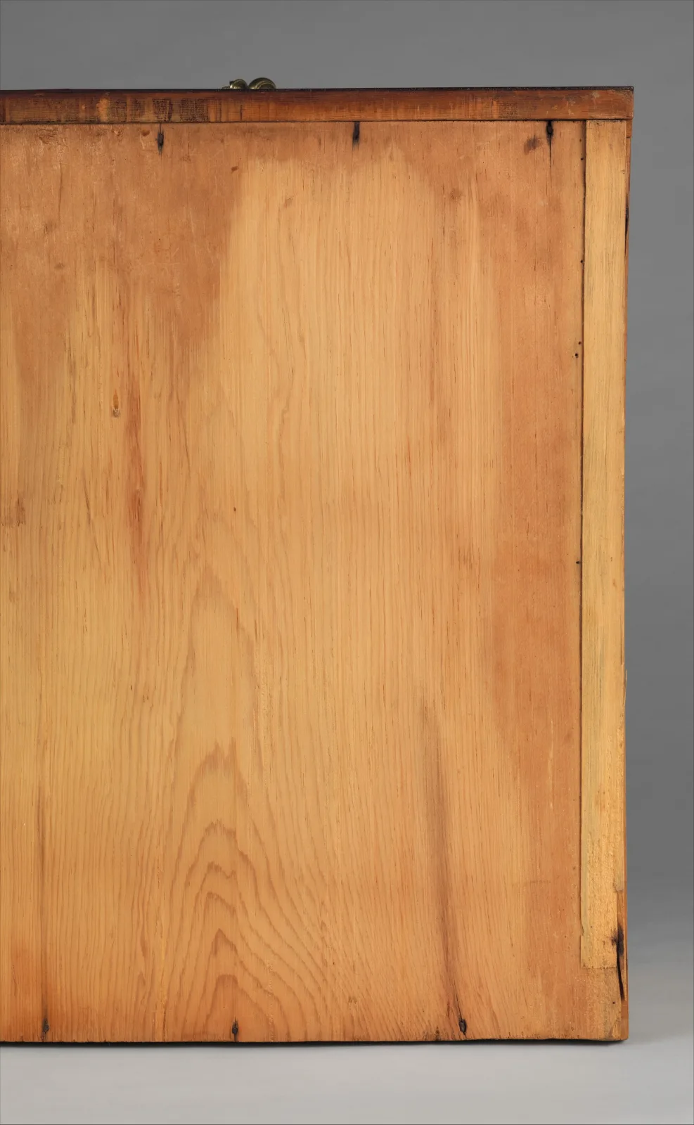 Underside of a pine drawer bottom with a brass pull visible at the top edge, the wood toned in uneven patches, darker toward the left and the ends, with old nail holes along the front