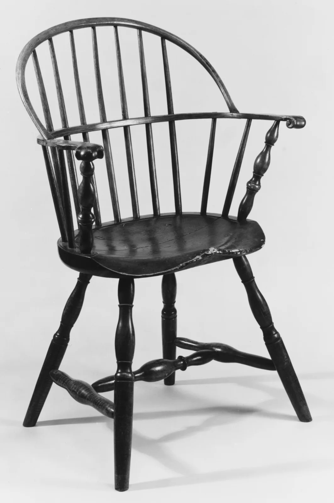 Black and white photograph of a painted sack-back Windsor armchair with turned legs and stretchers, the paint lost along the front edge of the seat and worn lighter on the arm rail