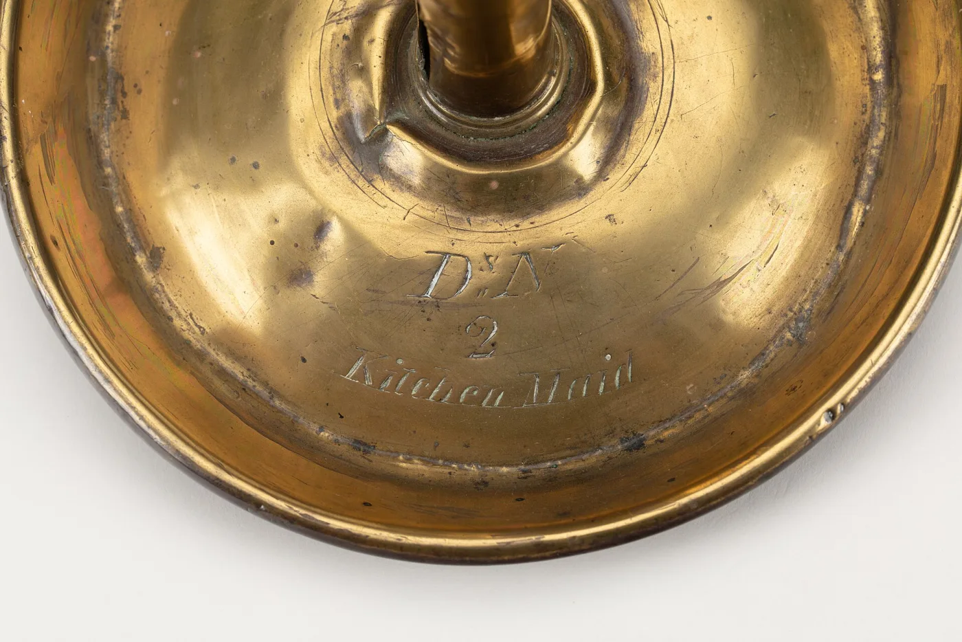 Underside of the dish-shaped base of a brass chamberstick, the bright yellow surface showing concentric turning rings and an engraved inscription reading D s N, 2, Kitchen Maid