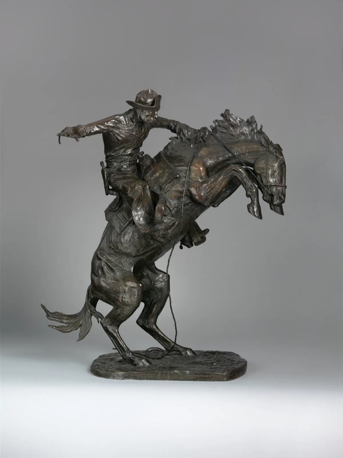 Dark patinated bronze of a cowboy on a rearing bronco, the horse balanced on its hind legs and the rider leaning back with hat raised, on a shaped naturalistic base