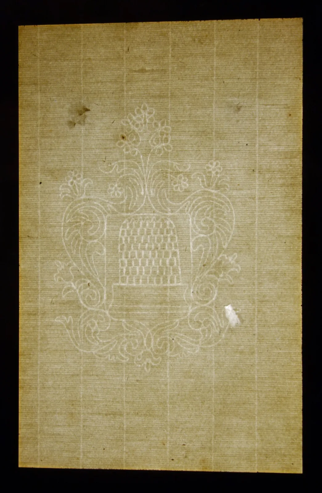 Aged and foxed laid paper lit from behind, showing a watermark of a beehive within scrolling rococo foliage, with laid and chain lines running through it, insect damage at the top and a small paper repair at the right