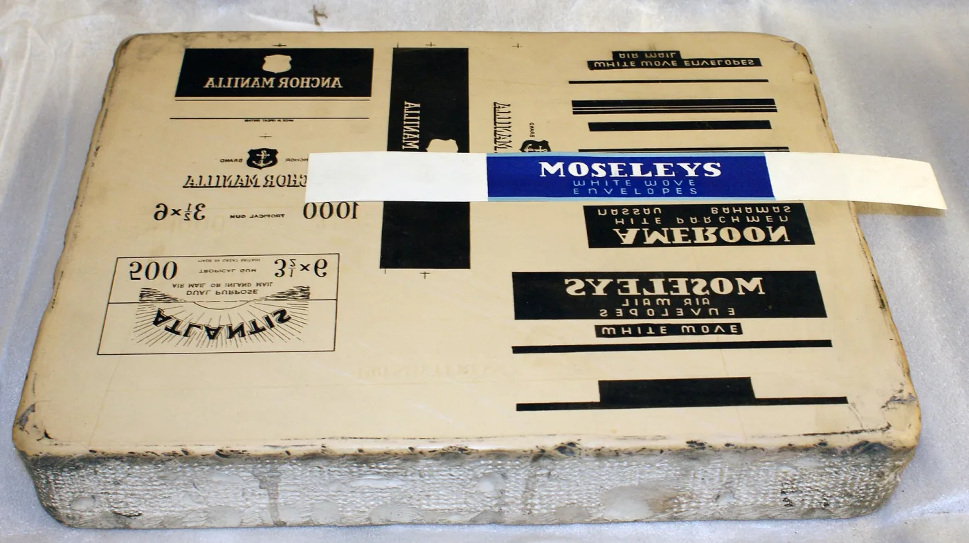 Thick slab of pale limestone with several commercial label designs drawn on its flat upper face in reverse, a printed blue and white envelope label laid across it showing the same design the right way round