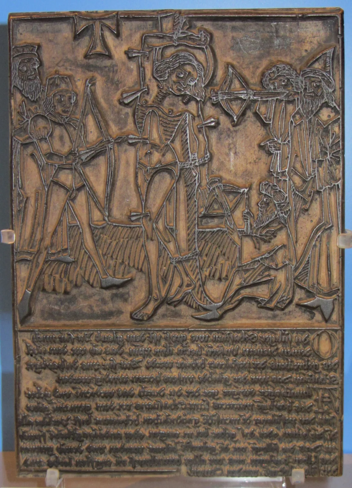 Rectangular wooden printing block carved in relief with archers shooting a bound saint, and eight lines of German text below, all of it cut in mirror image, standing on a museum mount