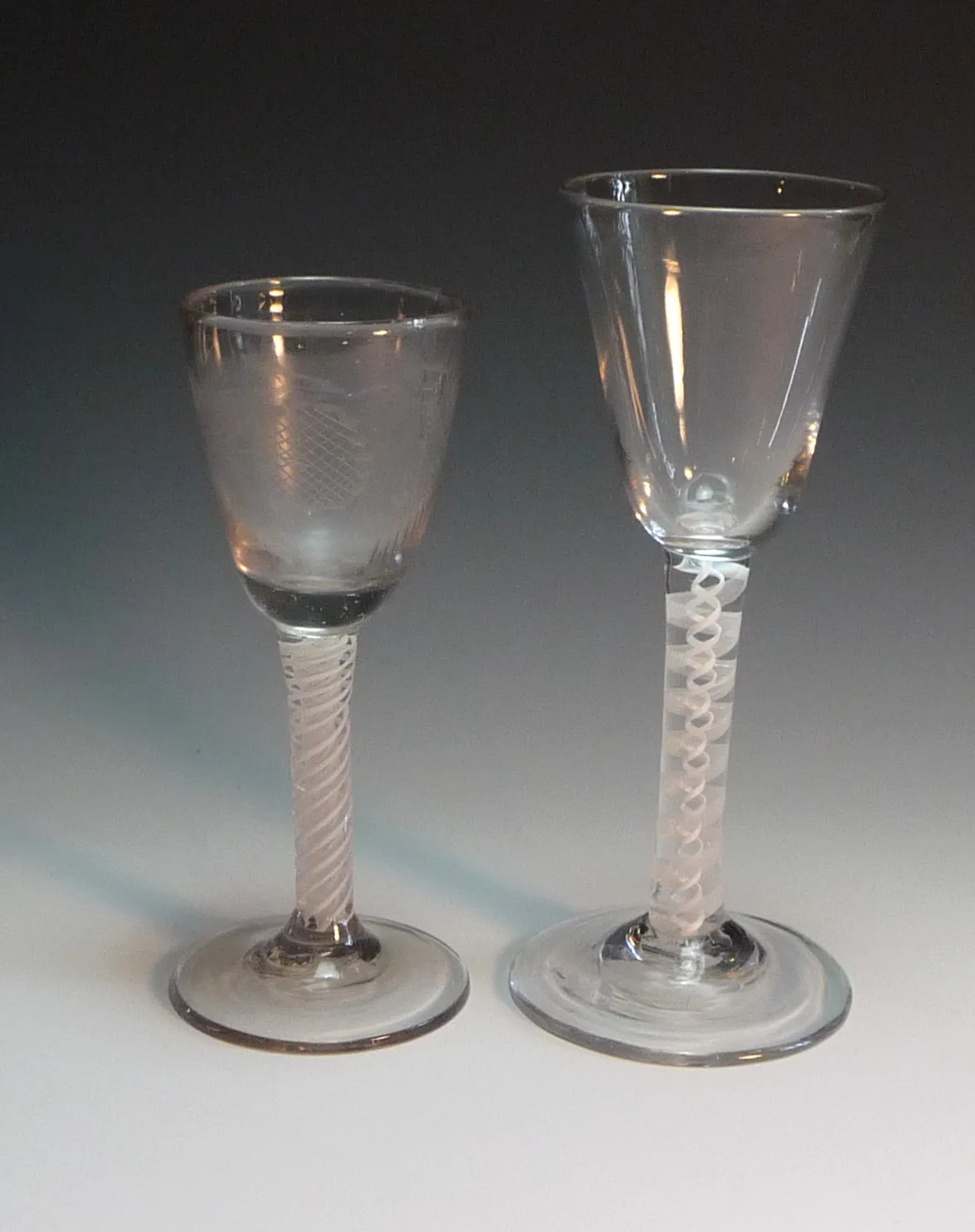 Two English opaque white twist wine glasses side by side, the left with a small engraved ogee bowl and a fine twist, the right with a larger bowl and a coarser more open twist