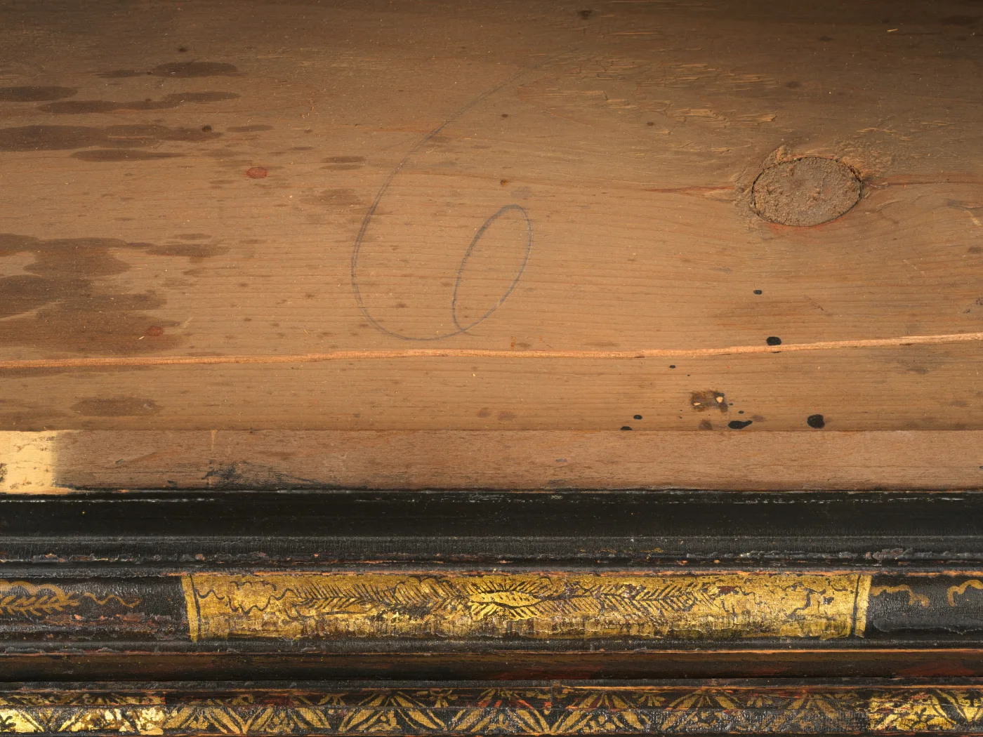 A pale pine board inside a case with a large numeral scribed in graphite, above a black japanned moulding with gilded decoration