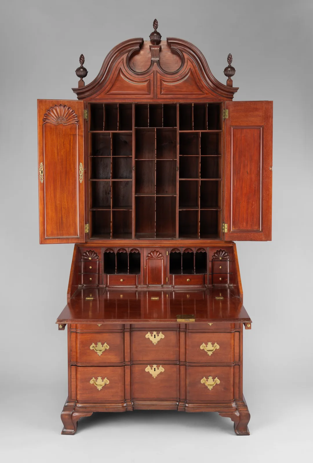 A mahogany desk and bookcase with its bookcase doors open to show pigeonholes, carved shells on the doors, and a slant-front desk open below